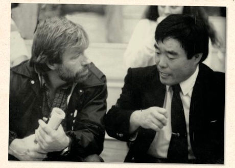 Two men in suits engaged in conversation, one holding papers. Background shows blurred people in seats. The mood is focused and attentive.
