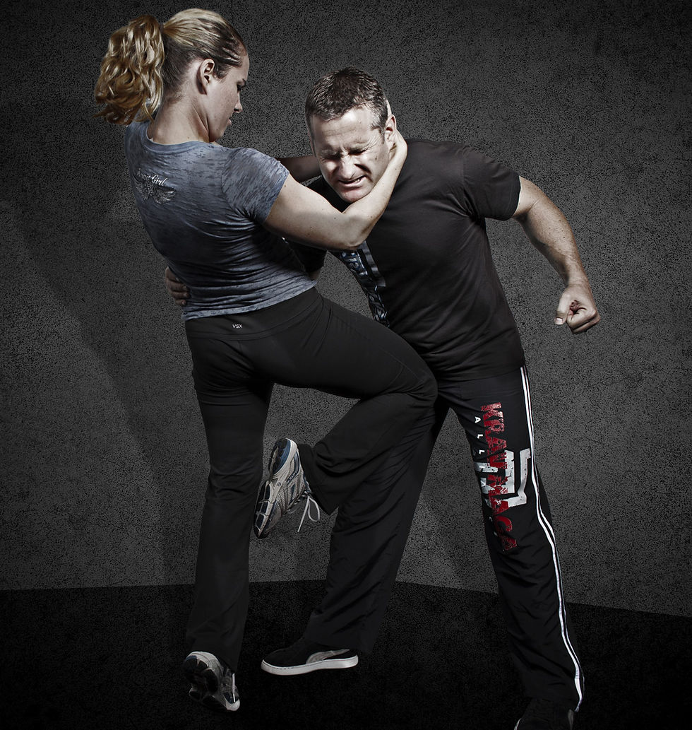 Woman in gray shirt performs a self-defense move on a man in black attire. Text on his pants reads Krav Maga. Intense mood, dark background.