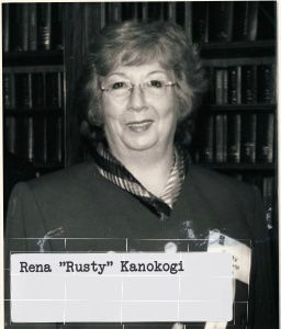 Smiling woman with glasses in front of bookshelves, wearing a dark jacket with patterned collar. Text reads "Rena 'Rusty' Kanokogi."