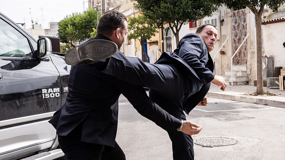 Two men in suits fight near a parked black Ram 1500 truck on a city street. One man kicks; intense action and focused expressions.