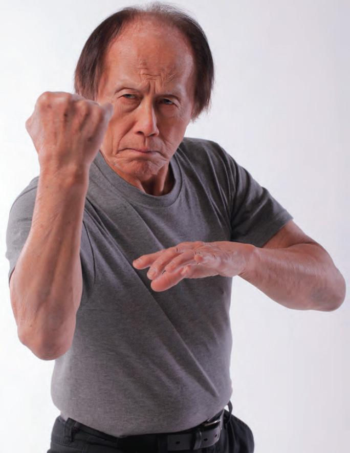 Man in gray shirt striking martial arts pose with fist raised, determined expression, white background. Focused and intense mood.