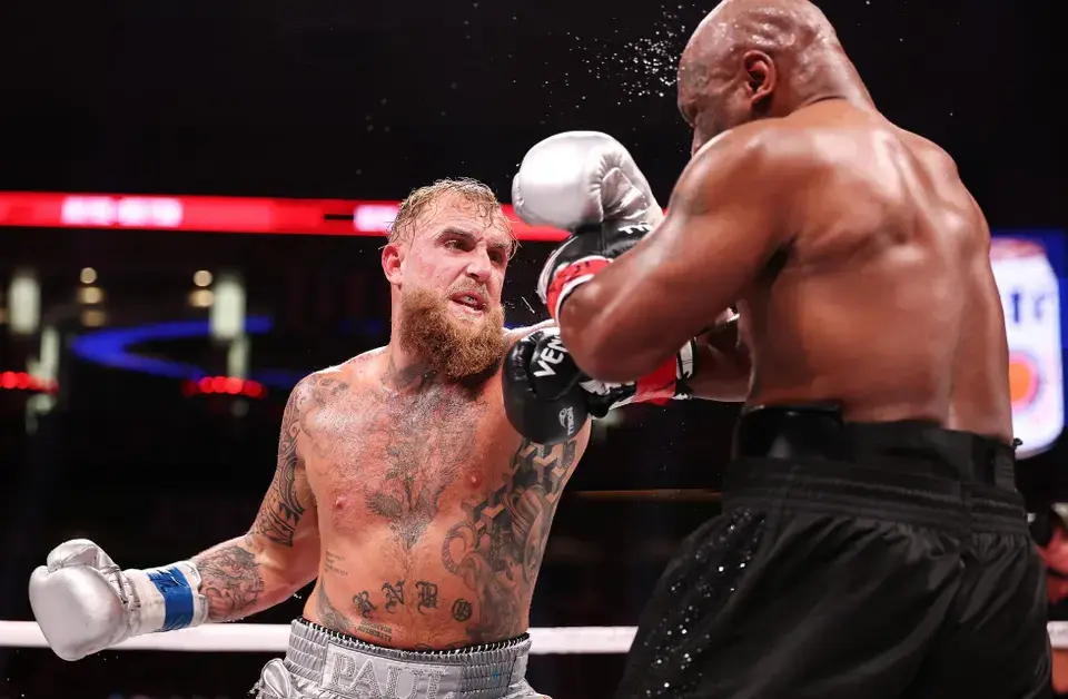 Two boxers in action; one punches the other in a ring. Tattoos visible. Focused expressions, bright arena lights, intensity palpable.