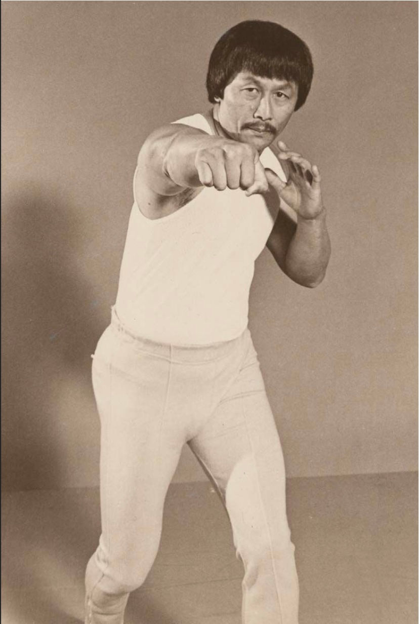 Martial artist in a white tank and pants strikes a fighting pose in a neutral indoor setting. Determined expression, sepia tone.