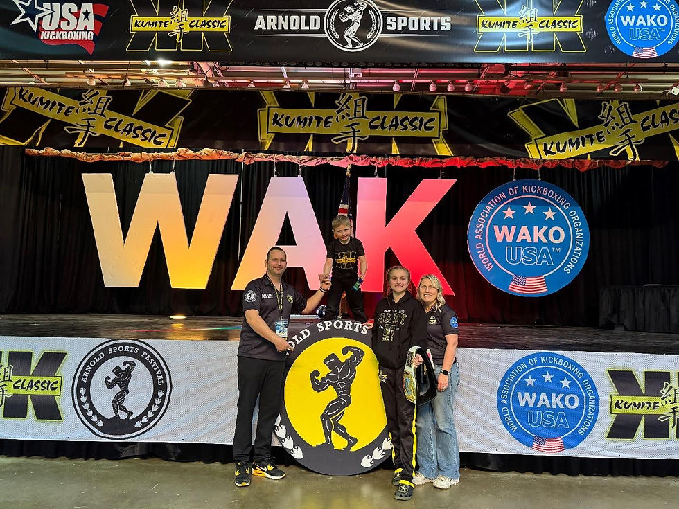 Four people smiling in front of a stage with "WAKO USA" and "Kumite Classic" banners. One child stands on a letter. Energetic mood.