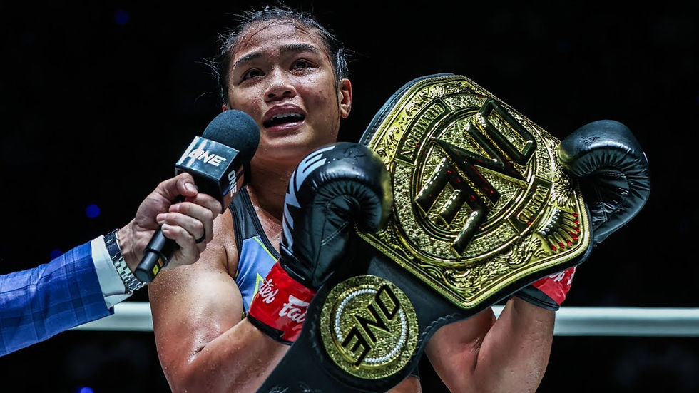 A boxer holding a gold championship belt in a ring, looks emotional. A microphone labeled "ONE" is held towards them. Dark background.