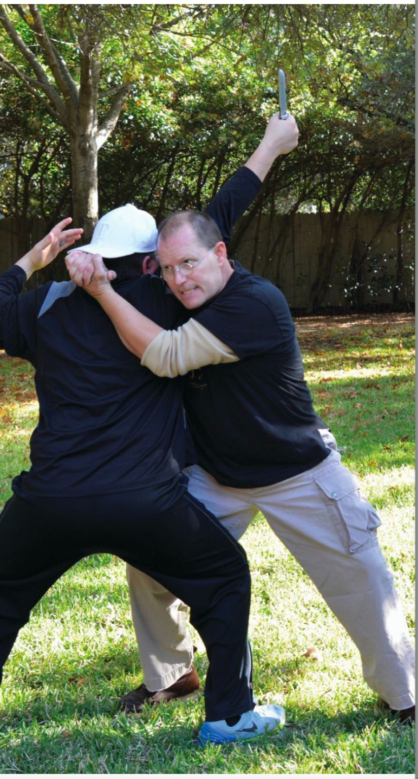 Two men are practicing self-defense in a grassy yard. One holds a knife visibly, while the other restrains him. Tension is evident.