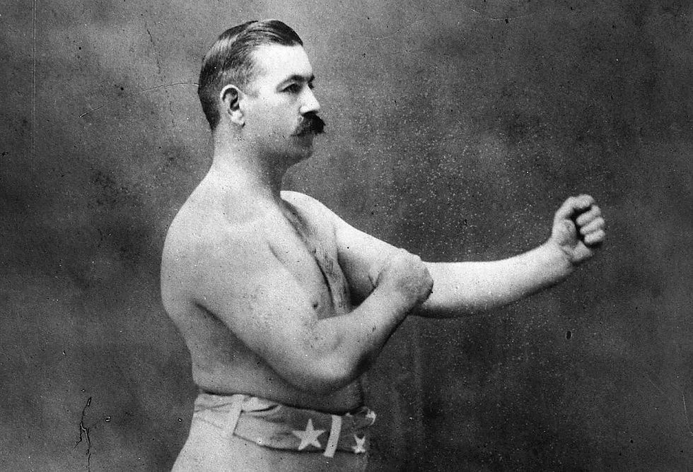 Man with a mustache in a boxing stance, bare-chested, wearing shorts with stars. Neutral background, vintage style photography.