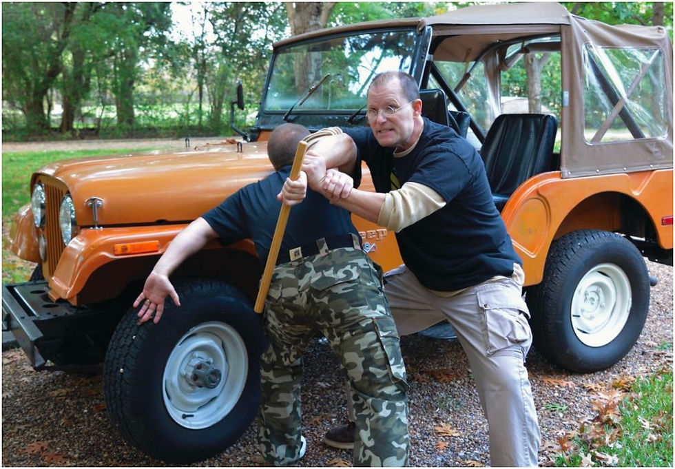 Two men practice martial arts with a stick next to an orange Jeep in a wooded area. One wears camo pants; the other, glasses.