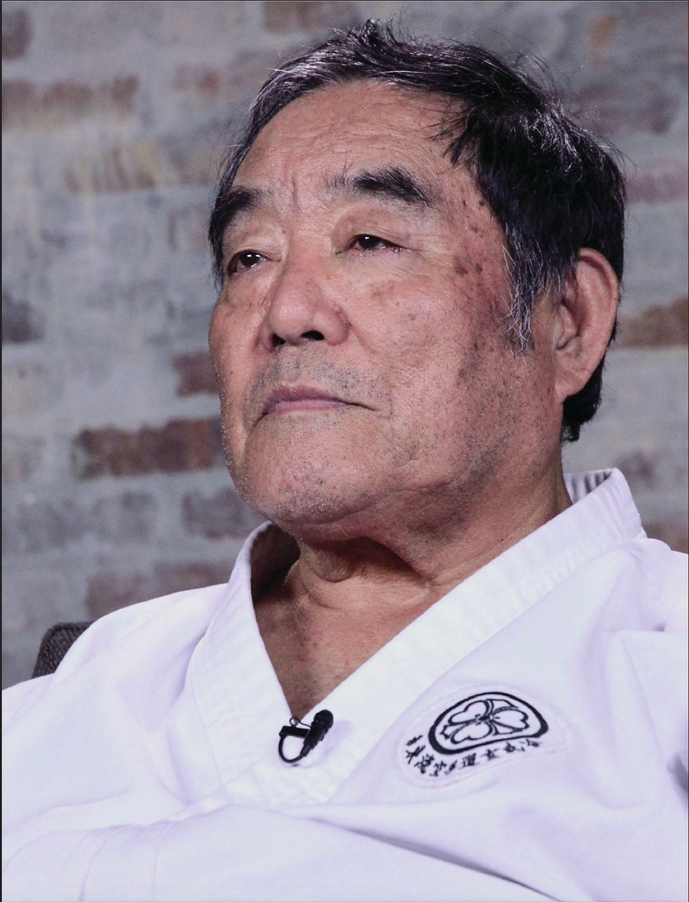 Elderly man in white martial arts uniform with emblem, looking thoughtful. Background features a blurred brick wall.