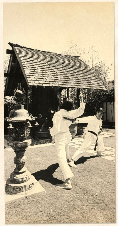 Two people in karate uniforms spar in a garden with a wooden structure and ornate lantern. Energetic atmosphere, black and white photo.