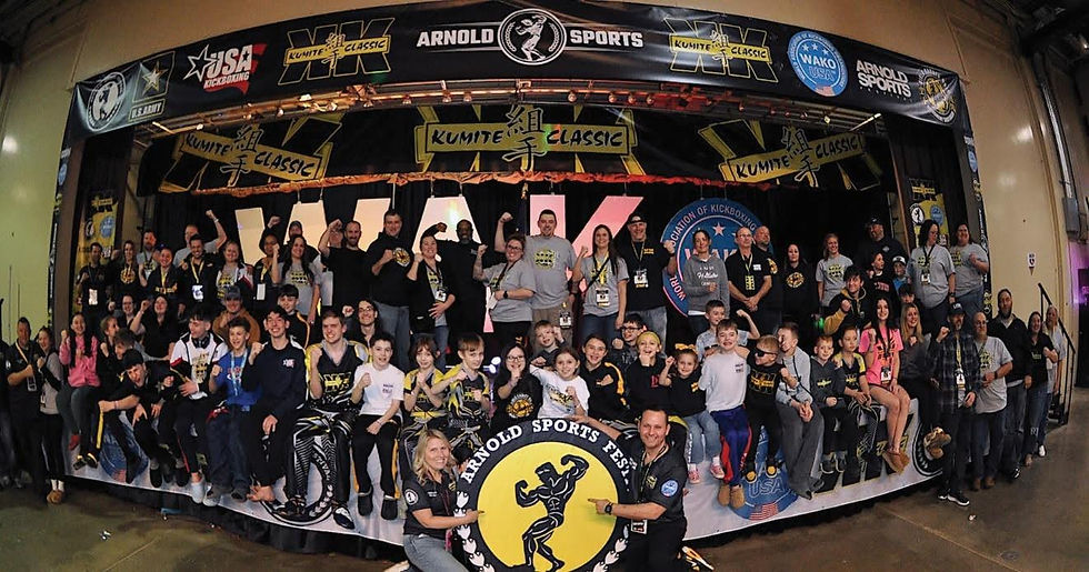 Large group on stage at a martial arts event, smiling and posing. Banners read "Kumite Classic" and "Arnold Sports." Energetic atmosphere.