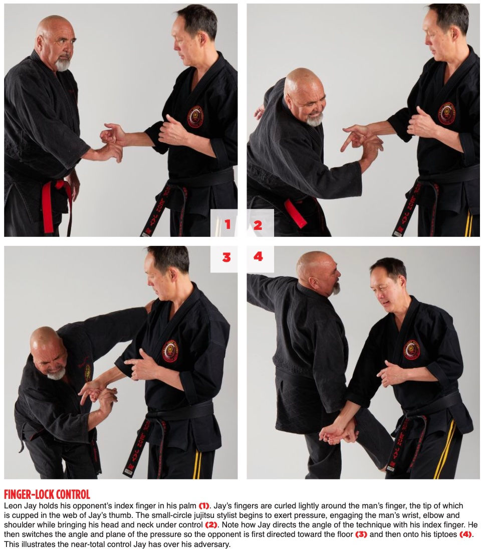 Two men in black martial arts uniforms demonstrate finger-lock control techniques. They're in a bright, neutral room, depicting focused action.