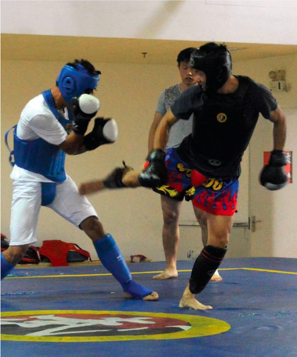 Two martial artists spar in a gym, wearing protective gear. One delivers a kick, while a third person observes. Intense action and focus.