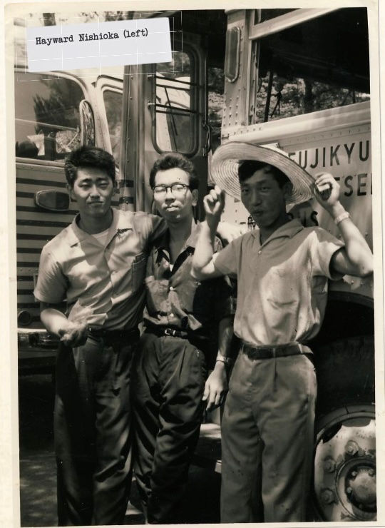 Three men pose in front of a bus, one wearing a straw hat. Partial text on the bus reads "FUJIKYU." The image is in black and white.