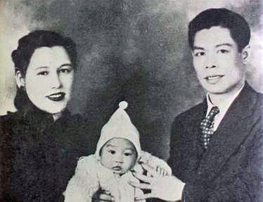 A man and woman hold a baby wearing a hat, sitting together with neutral expressions in a vintage black-and-white portrait.
