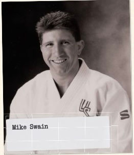 Smiling person in a judo uniform against a neutral background. Embroidery on the uniform. Text reads "Mike Swain" on a label.