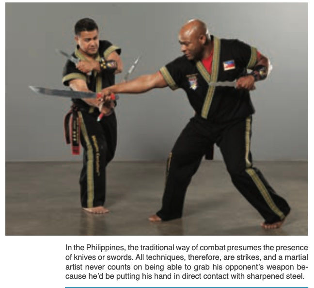 Two martial artists in black uniforms practice knife combat in a gray studio, blades crossed, with a Filipino flag patch visible.