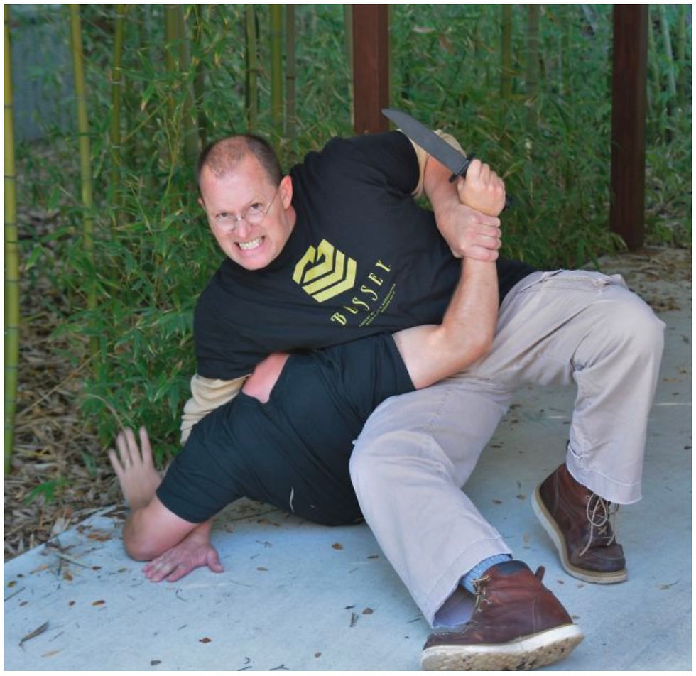 Man in a black t-shirt grapples with another on the ground, looking intense. Bamboo and cement visible in background, with knife in hand.