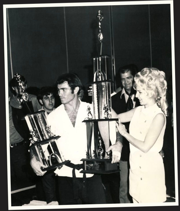 A man in martial arts attire holds two large trophies, assisted by a woman in a white dress. Smiling people stand in the background.