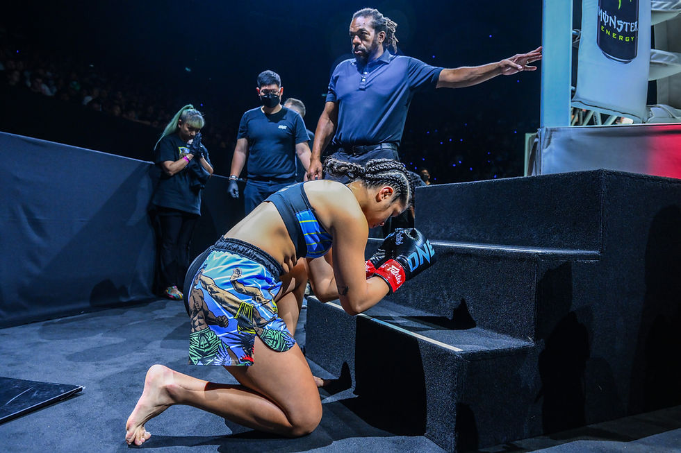 MMA fighter kneels in blue shorts and gloves, praying on steps. Referee points, two officials stand by. Dimly lit arena background.