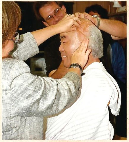 Man getting a bald cap fitted by two people in a workshop, smiling. The background is cluttered, creating a busy and joyful atmosphere.