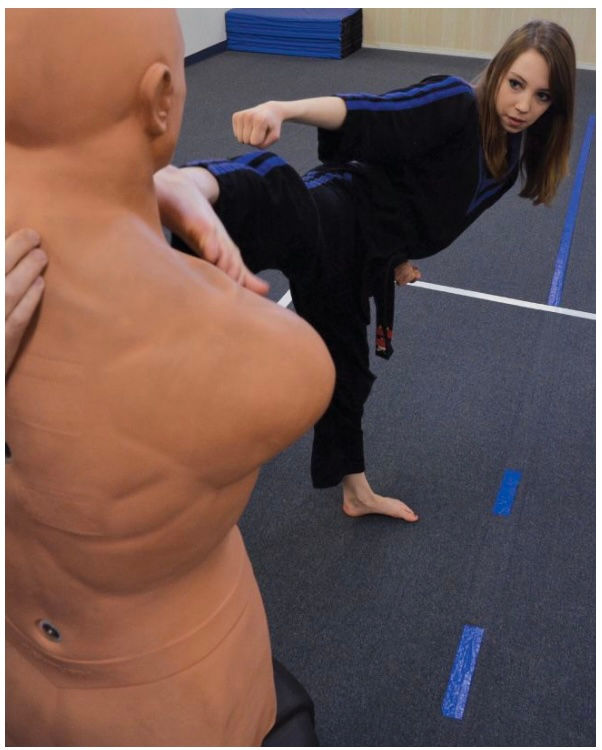 A woman in black martial arts attire kicks a dummy in a gym. Blue floor lines, focused expression, and workout mats in the background.