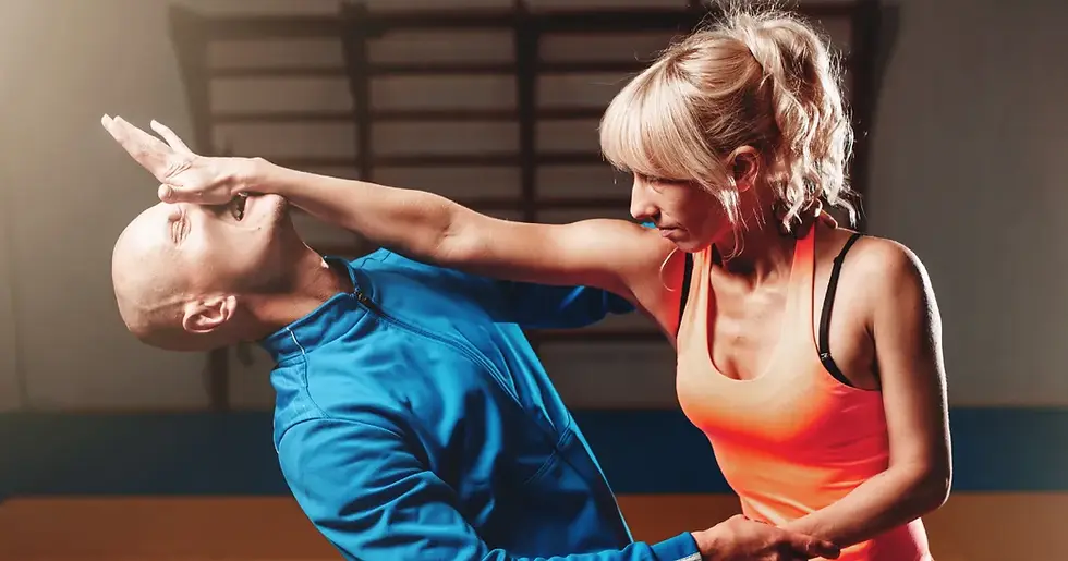 A woman in an orange tank top practices self-defense, pushing away a man in a blue jacket. They're in a gym setting, focused expressions.
