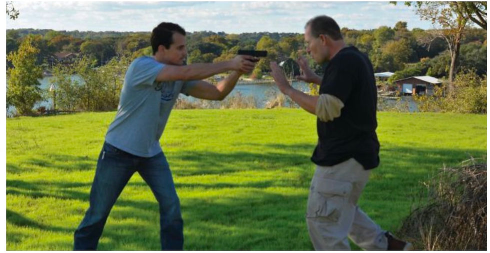 Two men in a grassy field; one aiming a gun, the other raising hands defensively. Background: lake and trees. Tense mood.