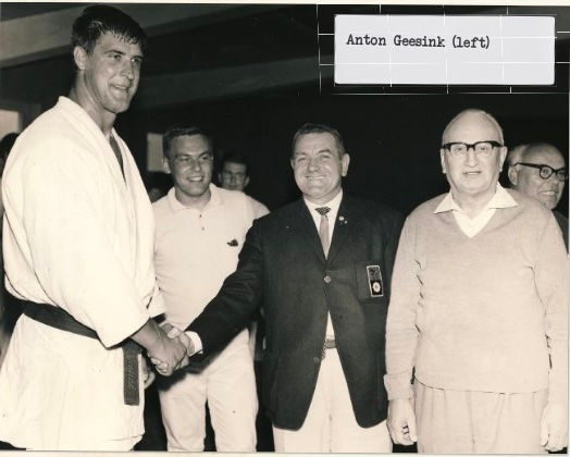 Four men in suits and a judo gi shake hands indoors. Black and white; text reads "Anton Geesink (left)." Smiling faces suggest a positive mood.