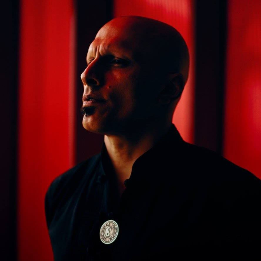 Bald person in profile with a meditative expression, wearing a black shirt with yin-yang symbol in a red-lit background.