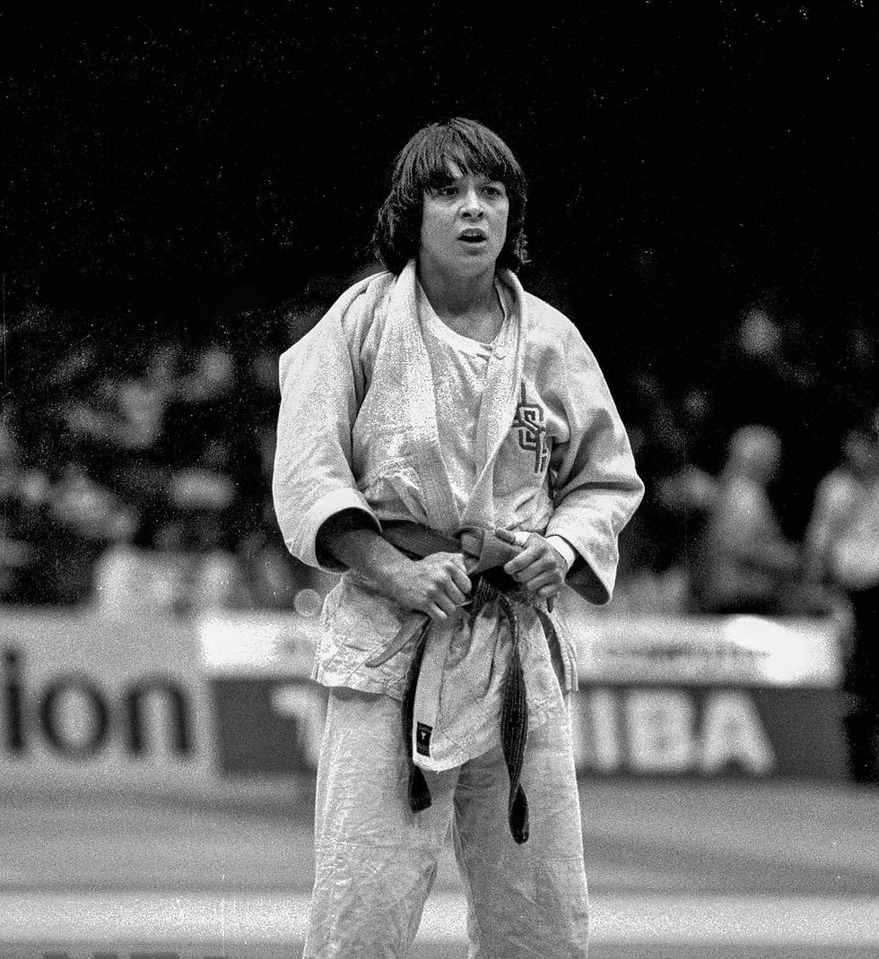 Judoka in a gi grips their belt in a stadium, with blurred spectators and signage in the background. Determined expression. Black and white.