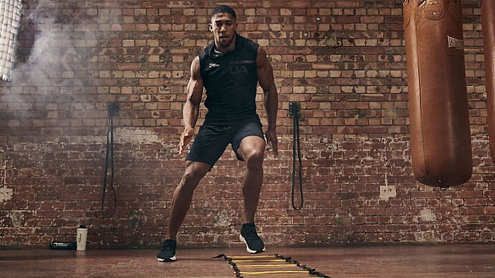 Man in black athletic gear performs ladder drills in a gym with exposed brick walls and punching bags. Determined expression, focused mood.