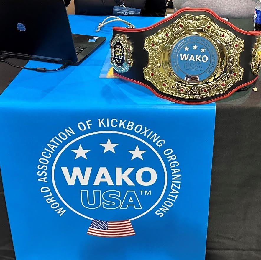 WAKO USA kickboxing belt on a blue table with logo; laptop nearby. Setting suggests a sporting event, with a focus on the championship belt.
