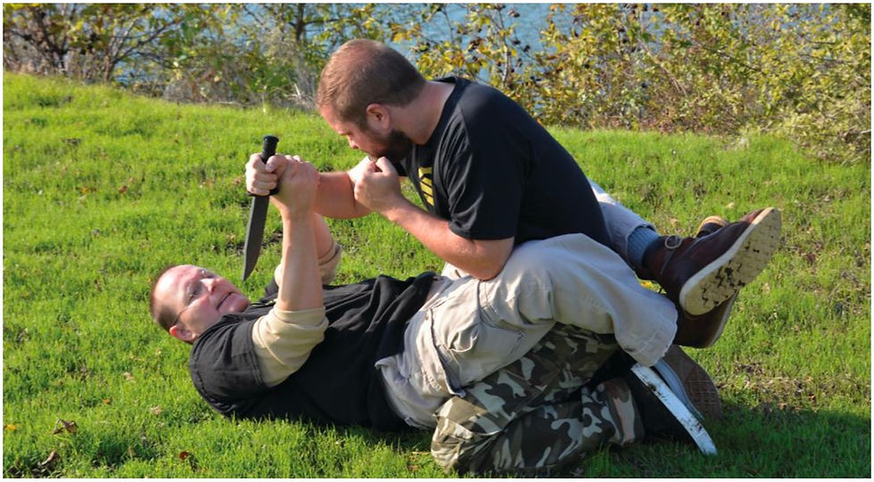 Two men in a grassy area; one on his back holding a knife, the other pinning him down. Intense struggle, surrounded by trees.