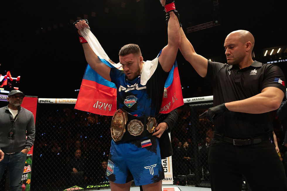 MMA fighter with championship belt and flag raised by referee in a dimly lit arena. Blue and red attire; audience watches in background.