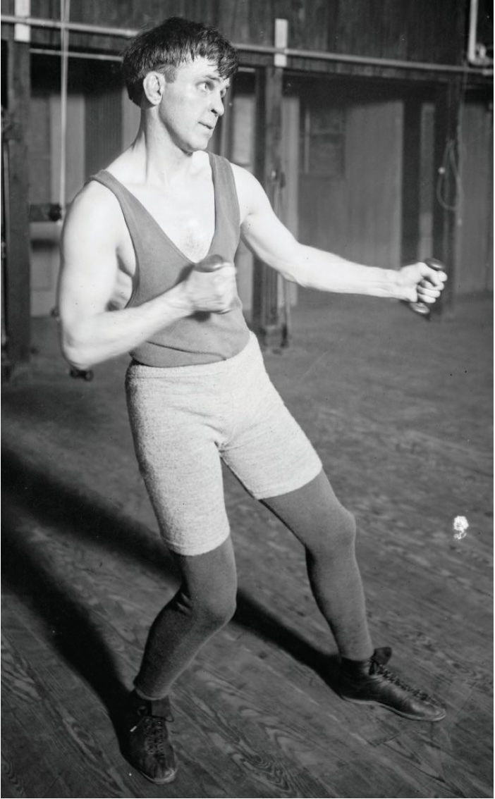 Man in workout attire boxing with dumbbells in a gym. Wearing a tank top, shorts, and tights. Focused expression, wooden floor visible.