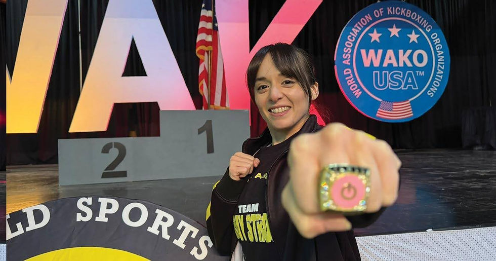 Smiling person shows a ring, standing before a podium. Background has large letters, USA flag, and WAKO sign. Energetic mood.