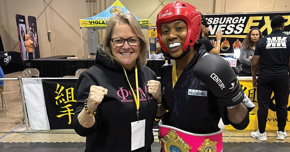 Woman in a black hoodie with raised fists stands next to a martial artist in red helmet and gloves. They smile at a sports expo.