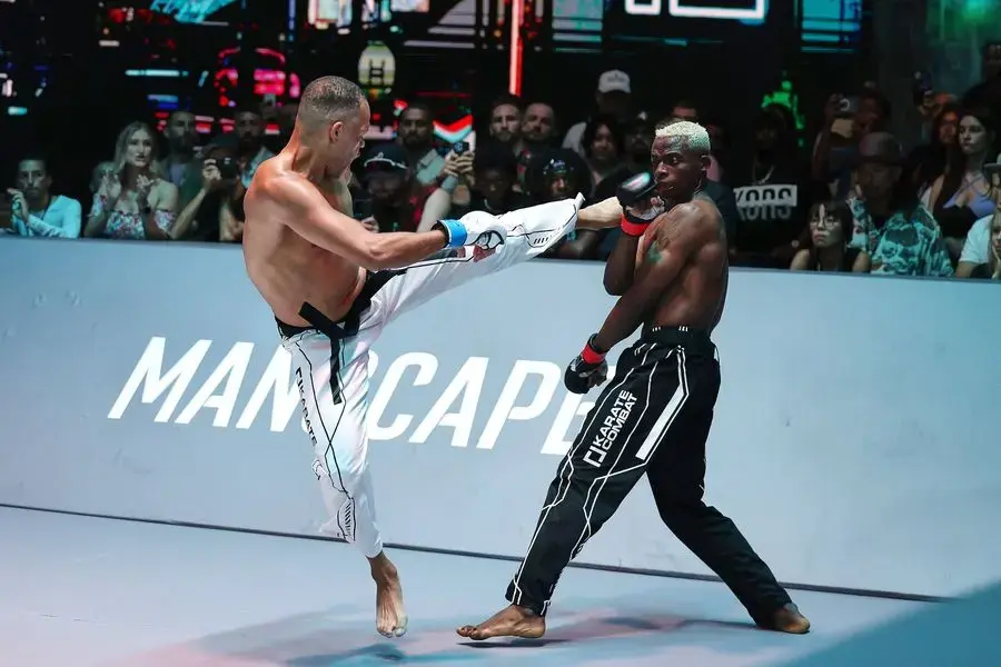 Two martial artists spar in a brightly lit arena. One delivers a high kick, while the other blocks. Intense focus, with spectators in the background.