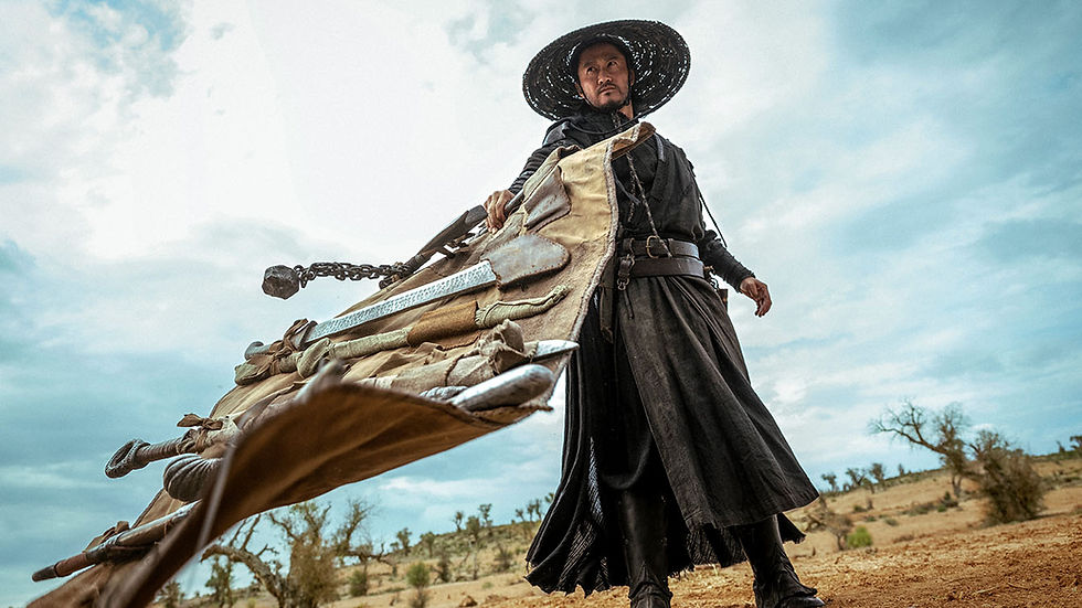 Man in black robes and straw hat stands in a desert, holding a huge chained weapon under a cloudy sky.