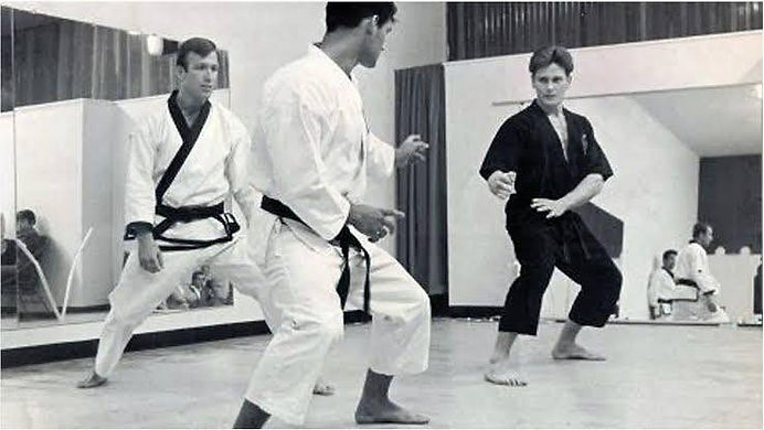 Three martial artists in gis practice stances in a mirrored studio, focused and ready.