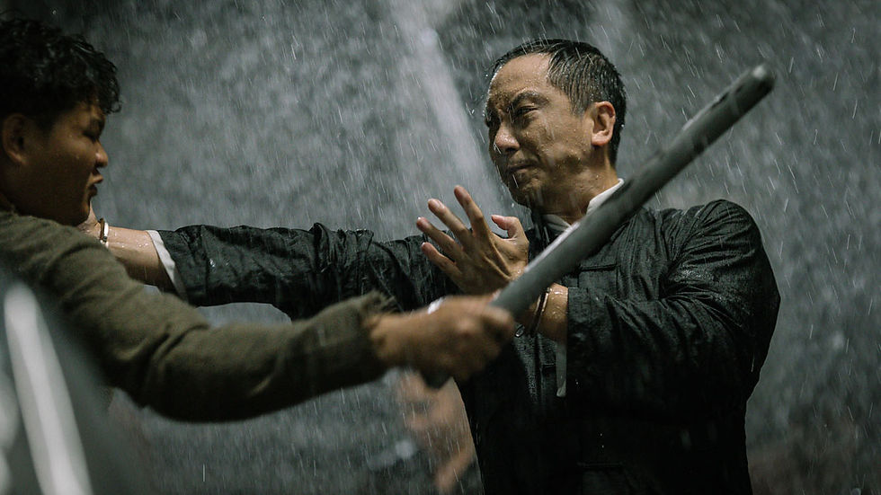 Two men fight with sticks in heavy rain, one in black blocking a strike; tense action scene.