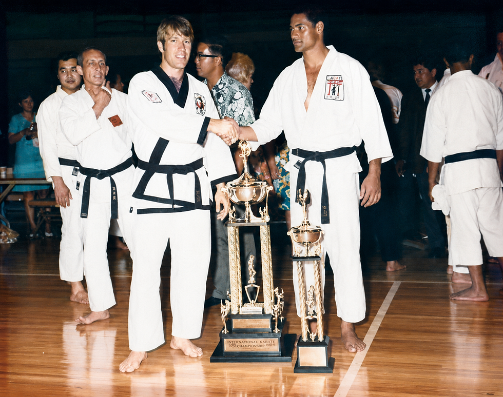 Two karate competitors in white uniforms shake hands beside tall trophies on a gym floor after a tournament, with spectators behind.