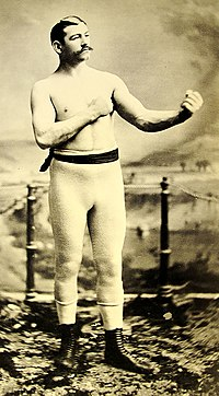 A man with a mustache poses in boxing stance, wearing boots and tights. Vintage sepia tone; background features a blurred landscape.