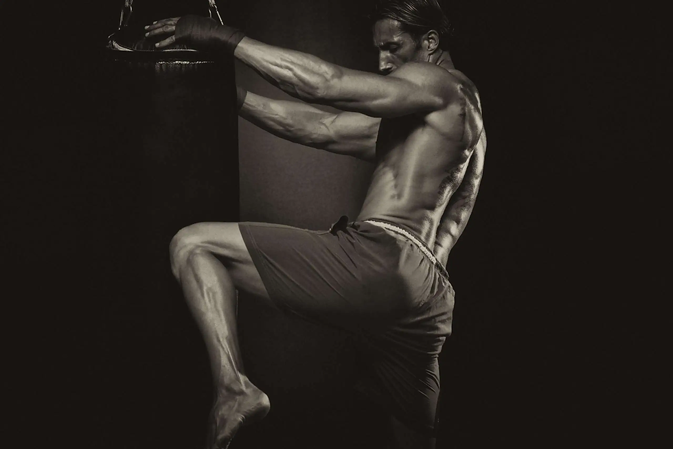Muscular man in shorts kneeing a punching bag in a dimly lit gym. Intense focus and energy. Monochrome tones.