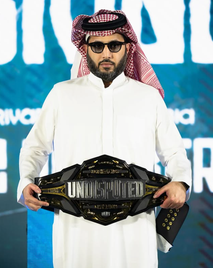 Man in a white robe and red headdress holds a black "UNDISPUTED" belt. Blue background with white text, serious expression.
