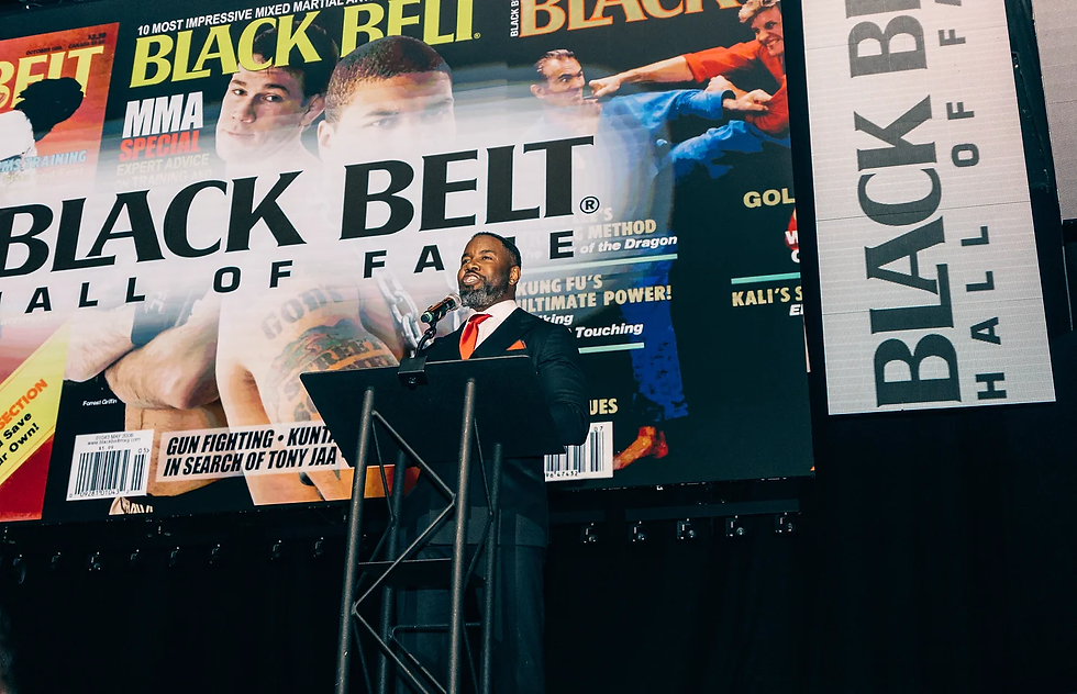 Man in suit speaks at a podium with "Black Belt Hall of Fame" backdrop, showing martial arts magazine covers. Mood is formal and celebratory.