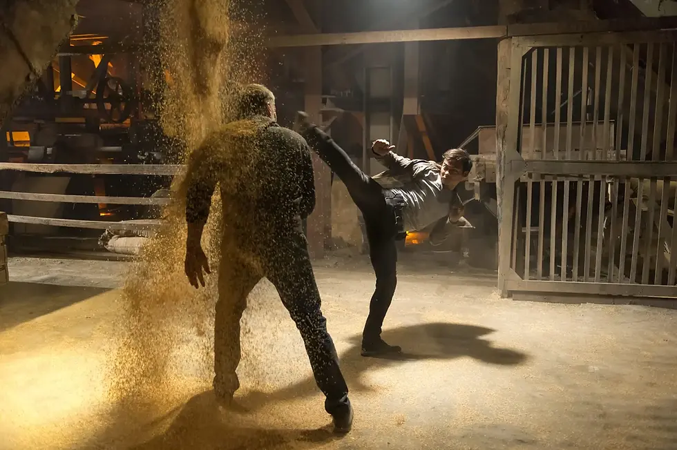 Man kicks another in a dusty warehouse, causing dust to rise. Industrial setting with metal bars and warm lighting. Energetic mood.