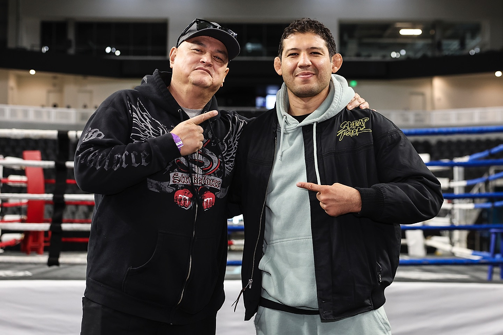 Two men in casual hoodies smile and point at each other in a boxing ring. One wears a "Strikeforce" and the other "SCRAP PACK" jacket.