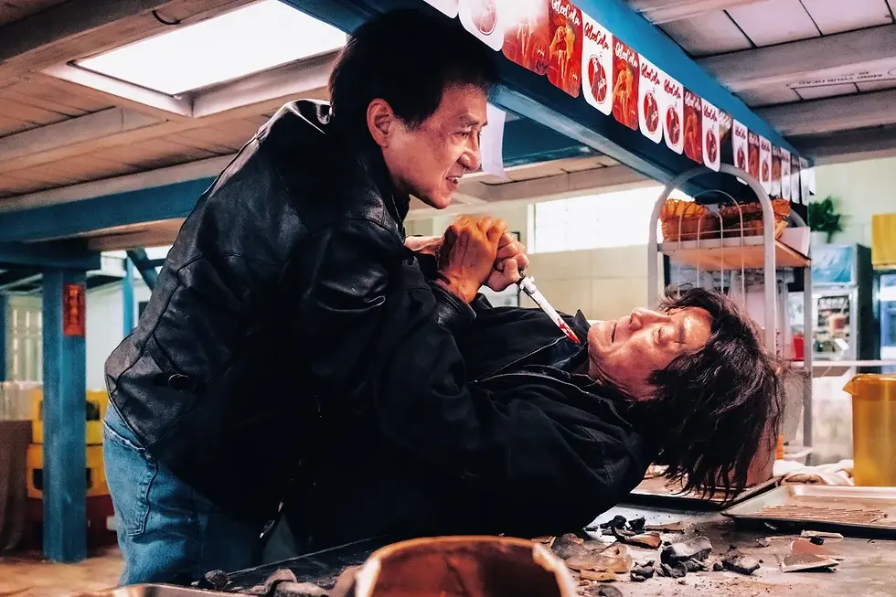 Two men in a tense struggle on a table with a knife between them, set in a brightly lit room with colorful posters above.